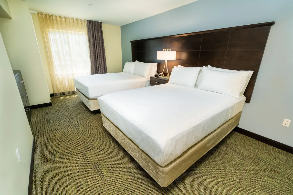 A spacious, tranquil bedroom featuring two plush Queen beds, a central nightstand with a lamp, and a large window dressed with blackout curtains. Sunlight streams through the window, highlighting the inviting, restful atmosphere of the room, ideal for families, friends, or colleagues.