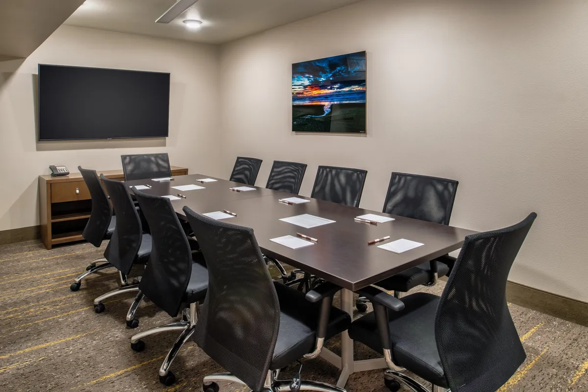 A modern conference room furnished with a large table, comfortable rolling chairs, and a TV screen for presentations. The space is designed for collaboration, with artwork on the wall providing a touch of sophistication. Colleagues are pictured in the midst of a meeting, suggesting productivity and teamwork.