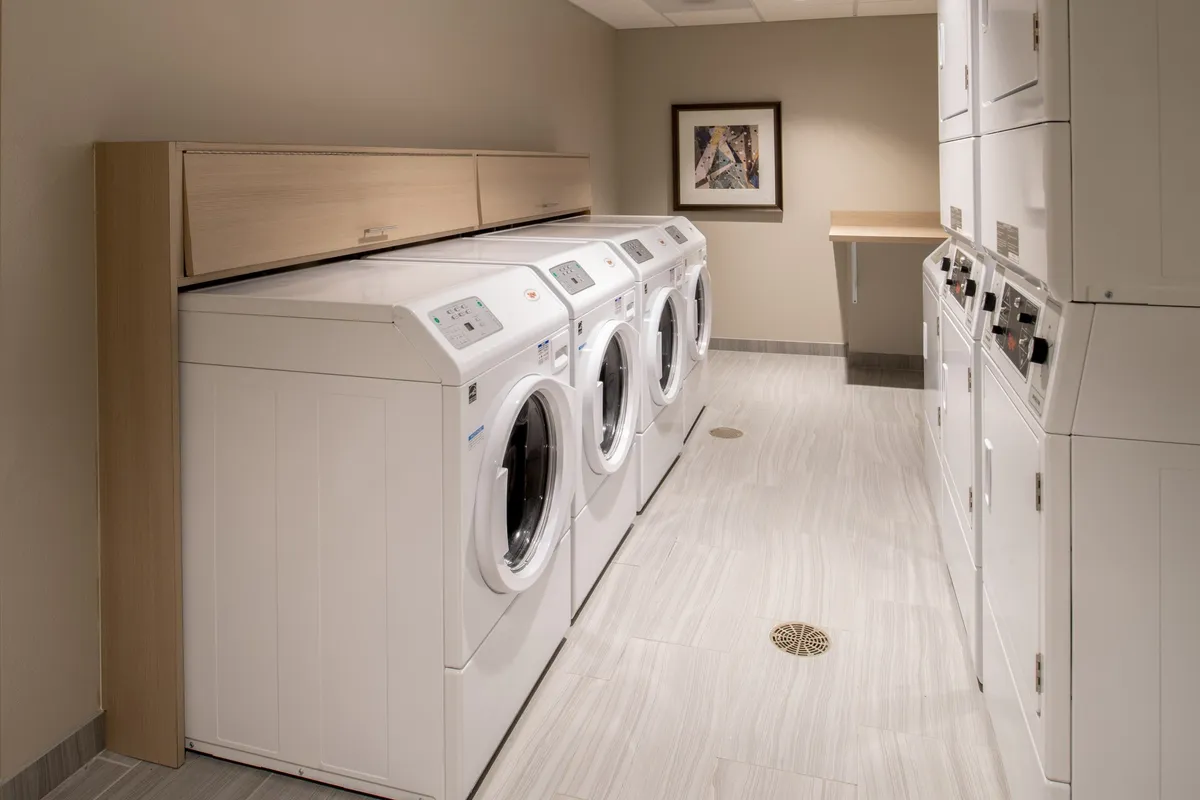 The self-serve laundry room is depicted with multiple washing machines and dryers, ample counter space for folding, and framed artwork, creating a clean, practical, and welcoming environment.