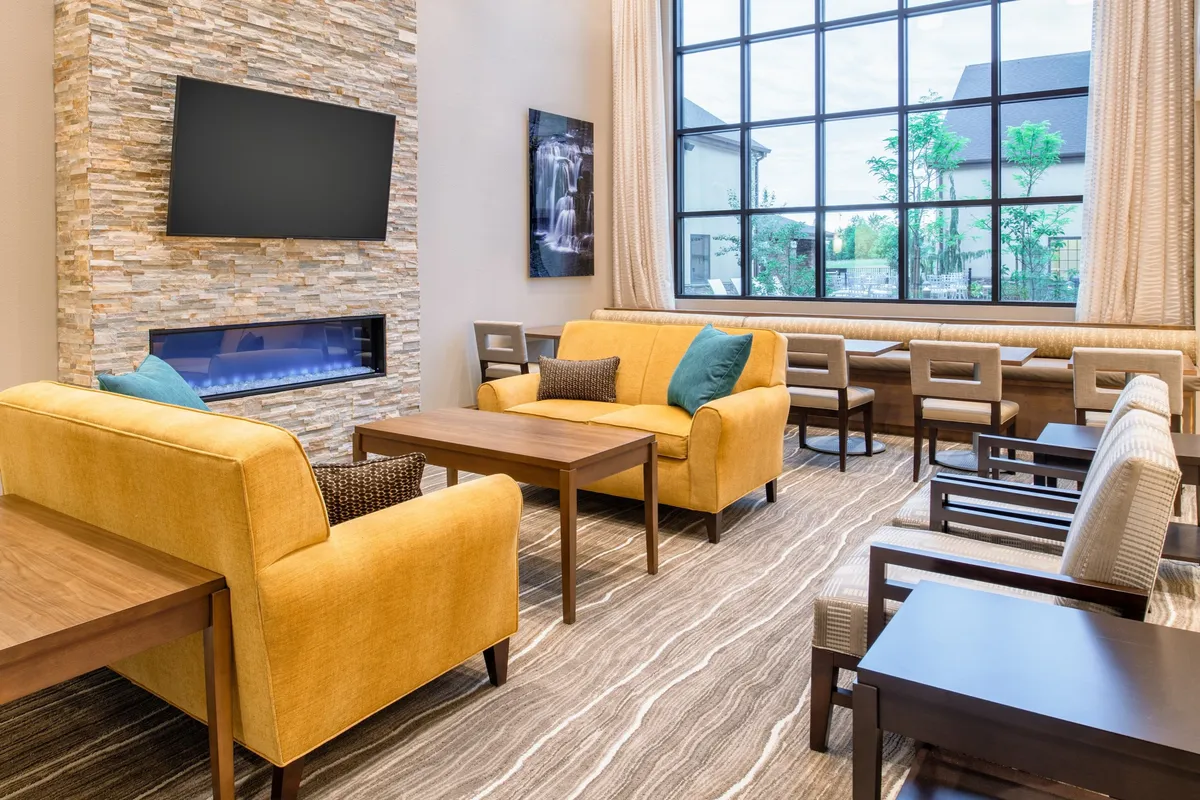 A warm, inviting living area with plush yellow sofas arranged around a coffee table, a large window letting in natural light, a stone wall with a mounted flatscreen TV above a modern fireplace, and tasteful artwork on the walls. The space exudes comfort, ideal for both relaxing and socializing.