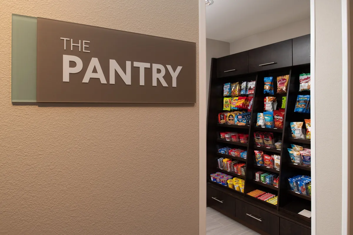 The image shows a welcoming pantry area within Staybridge Hillsboro North, featuring well-stocked shelves filled with a variety of snacks and beverages. The 'pantry' sign is visible, and guests are seen browsing or socializing, creating a casual and friendly atmosphere.