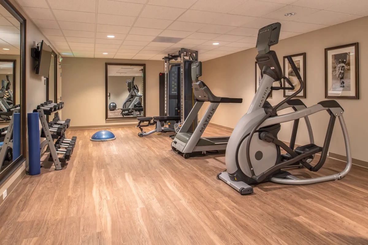 A well-equipped fitness center is shown, including an elliptical machine, weight bench, large mirrors, and an exercise ball. The space is bright and motivating, emphasizing opportunities for guests to work out or take group classes.