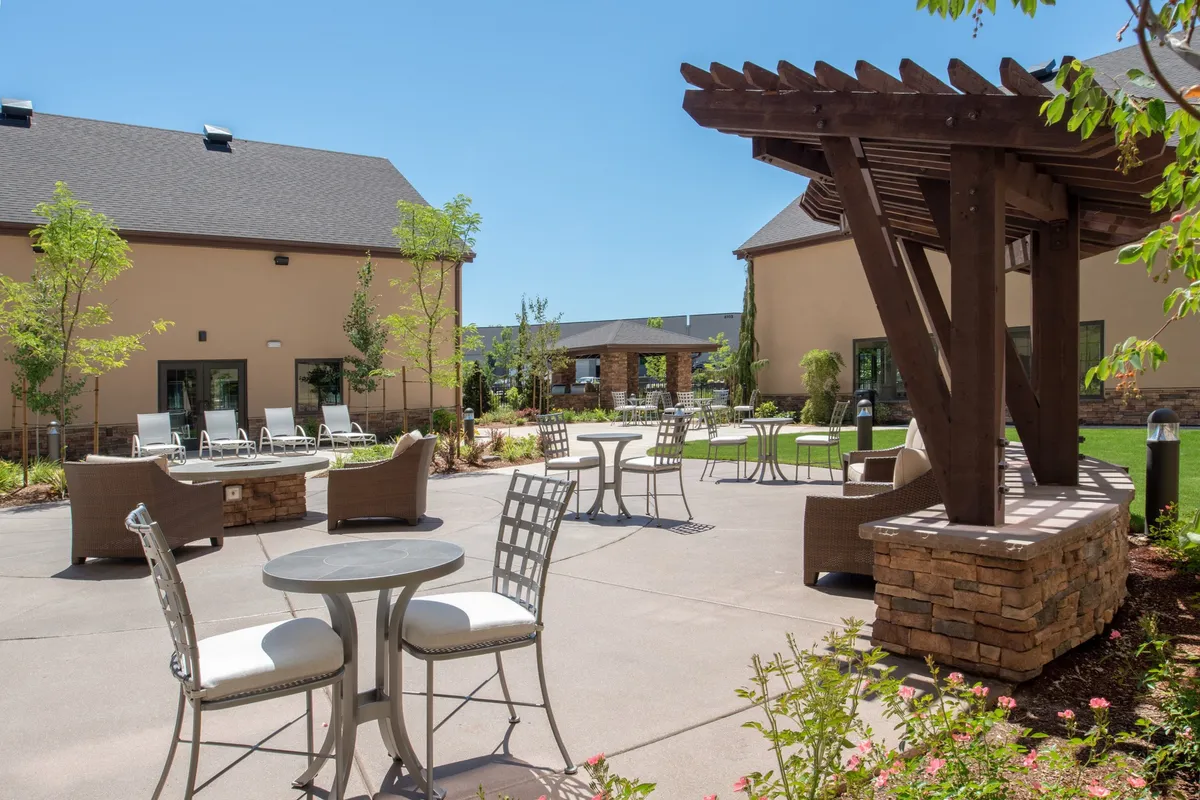The image shows an inviting outdoor seating area beneath a pergola, surrounded by lush greenery. The setup encourages relaxing, socializing, and outdoor dining, capturing the essence of shared spaces ideal for BBQs or gatherings. The tranquil, social mood is accentuated by comfortable furnishings and the natural setting, perfect for enjoying fresh air meals.