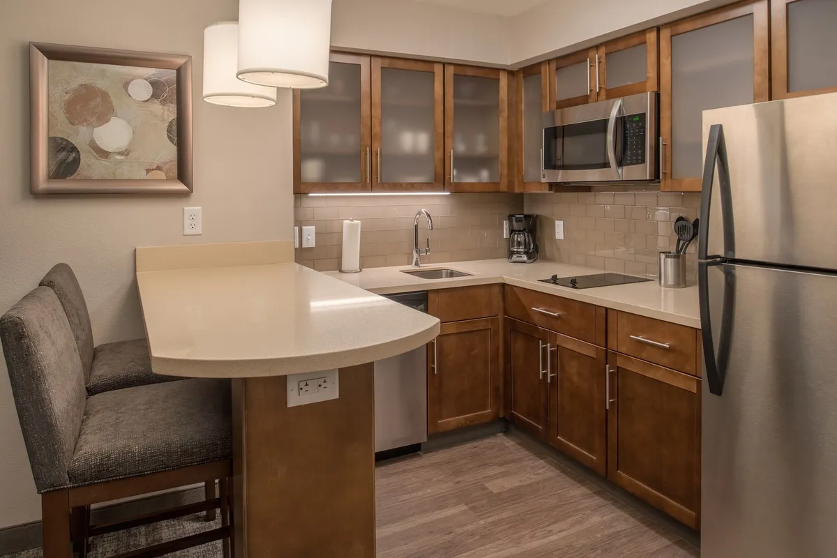 This cozy, contemporary kitchen interior features a stylish bar counter, modern kitchen cabinets, and prominently displayed appliances. The space is arranged for both cooking and socializing, with the bar creating a natural hub for casual dining or conversation. Subtle artwork and soft lighting contribute to the comfortable atmosphere, making it inviting for preparing meals or gathering with friends.