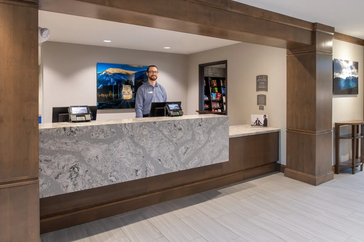 The image shows the reception desk at Staybridge Hillsboro North, with a staff member welcoming guests. The reception area is bright and inviting, featuring artwork that hints at the local culture, setting a professional yet welcoming tone for business travelers arriving at the hotel.